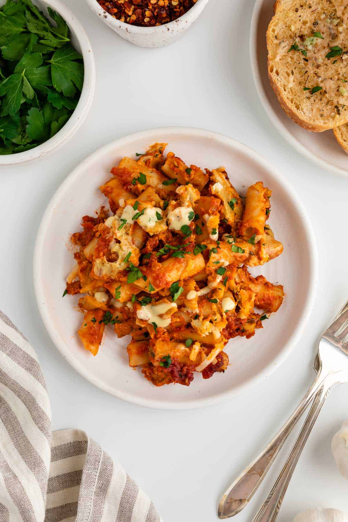 a serving of vegan baked ziti on a white plate surrounded by a striped linen napkin, two forks, a bowl of fresh parsley, a bowl of crushed red pepper, and vegan garlic bread