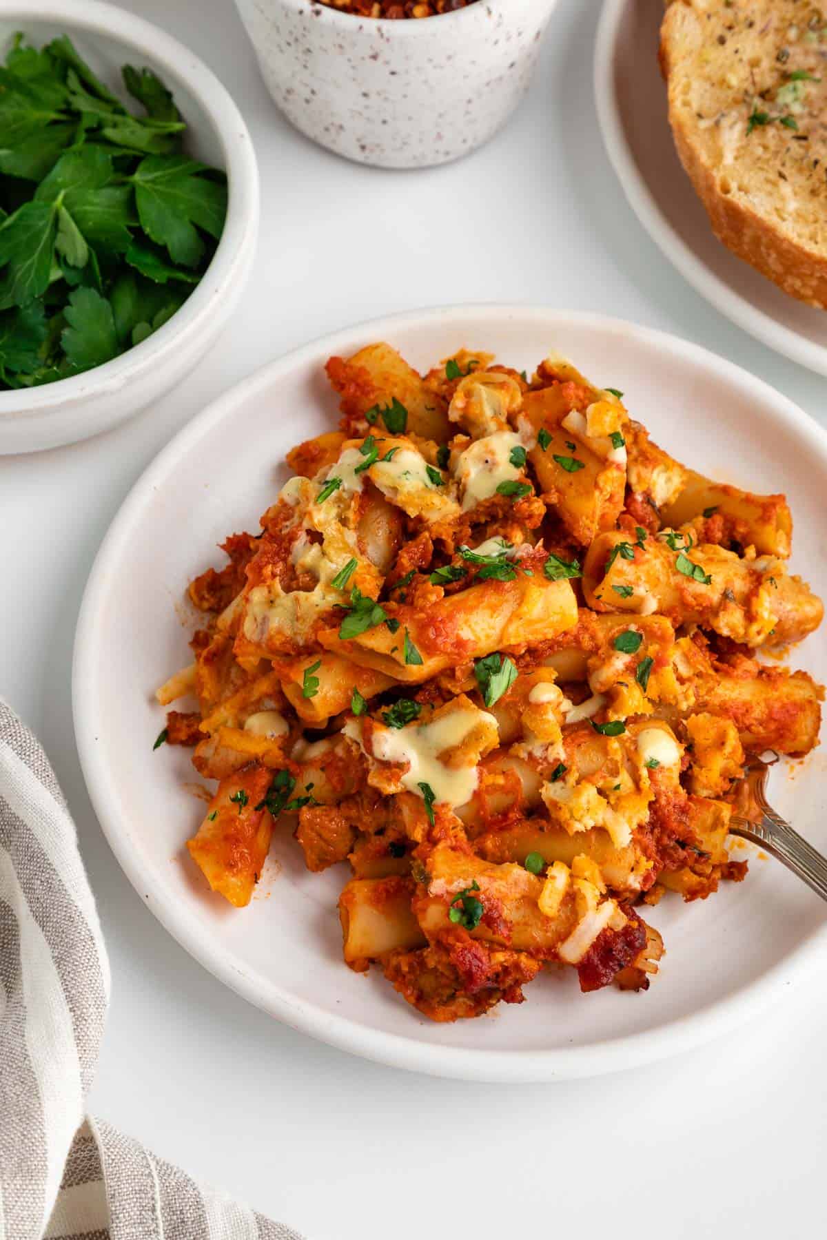 a serving of vegan baked ziti on a white plate beside a striped linen napkin, bowl of parsley, and vegan garlic bread