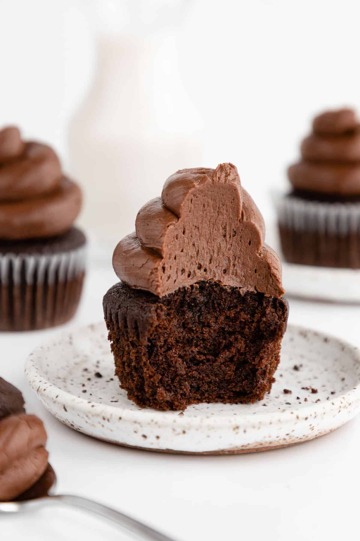 a partially eaten vegan chocolate cupcake with chocolate buttercream frosting sitting on a ceramic dessert plate, surrounded by a glass of almond milk and additional cupcakes
