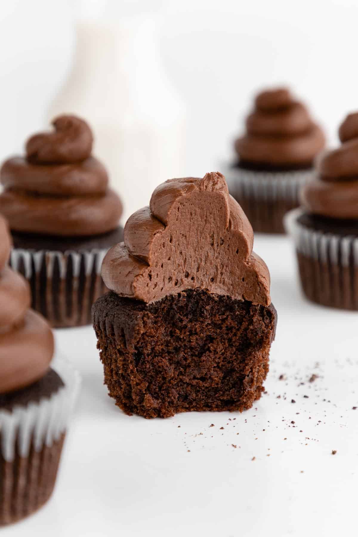 a partially bitten vegan chocolate cupcake with chocolate buttercream frosting, surrounded by more cupcakes