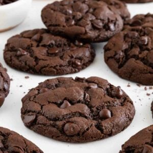 vegan double chocolate chip cookies on a white surface beside a bowl of cocoa powder and another bowl of chocolate chips