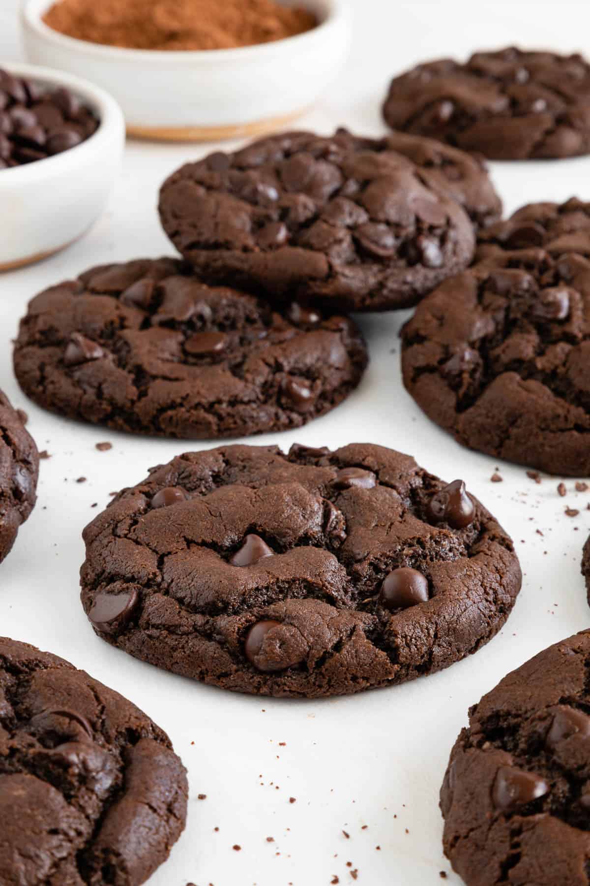 vegan double chocolate chip cookies on a white surface beside a bowl of cocoa powder and another bowl of chocolate chips