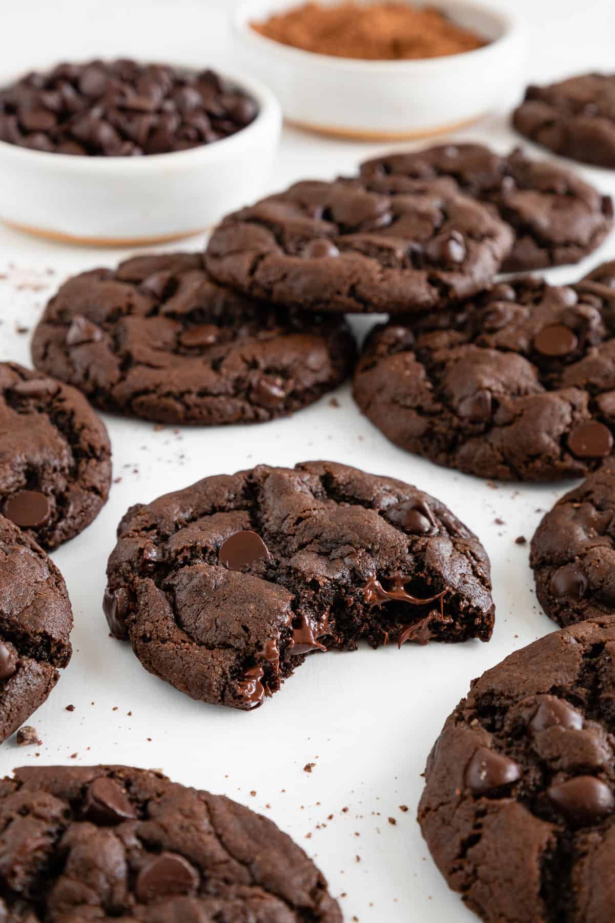 a gooey vegan double chocolate chip cookie with a bite taken out of it, surrounded by a dozen more cookies and a bowl of cocoa powder