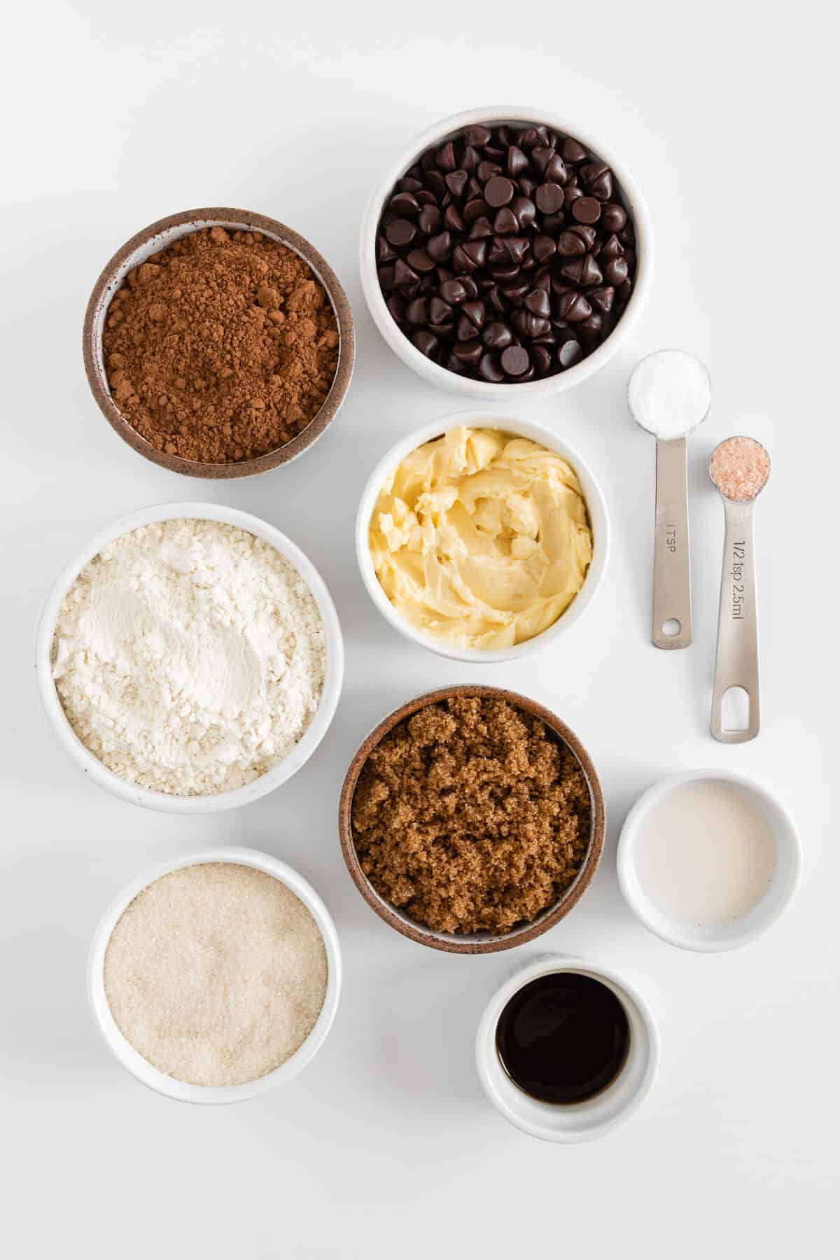 small white bowls filled with chocolate chips, cocoa powder, flour, vegan butter, sugar, brown sugar, vanilla extract, and non-dairy milk