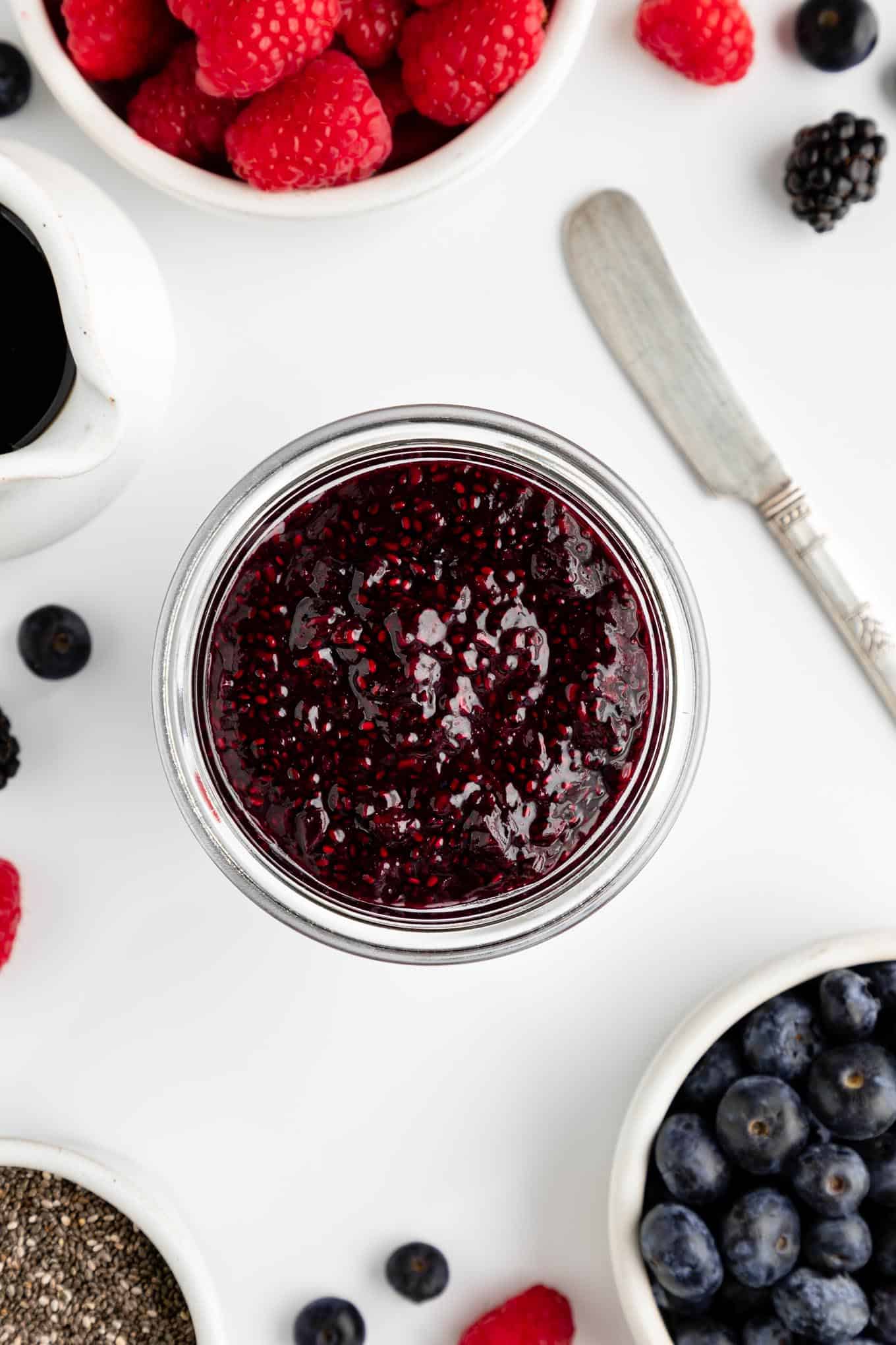overhead image of berry chia seed jam inside a glass weck jar surrounded by mixed berries and a knife