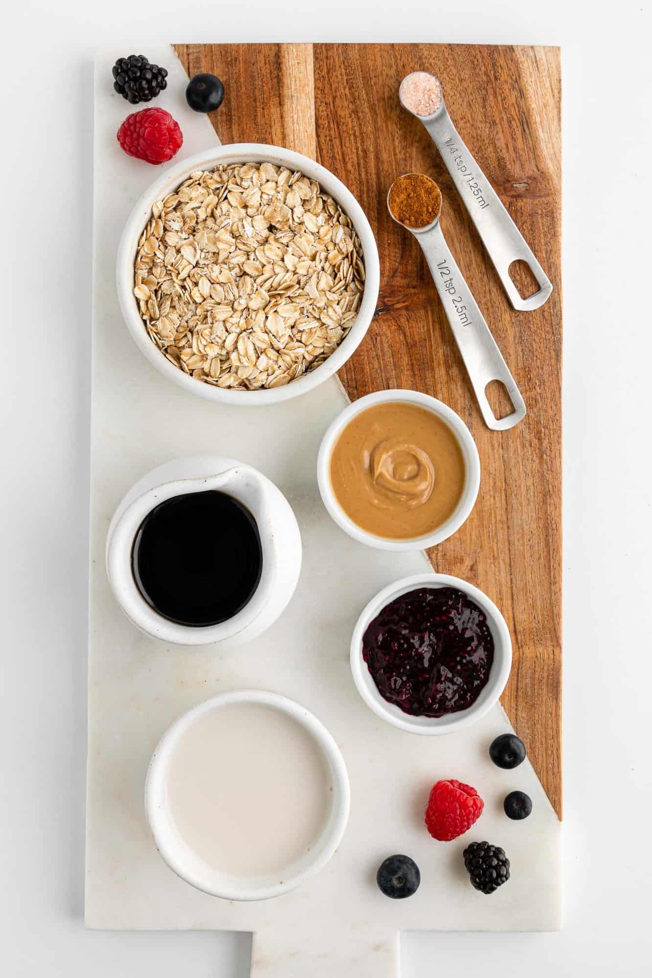 a wooden and marble serving board topped with ingredients for pb&j oatmeal, including oats, almond milk, maple syrup, and berry jam