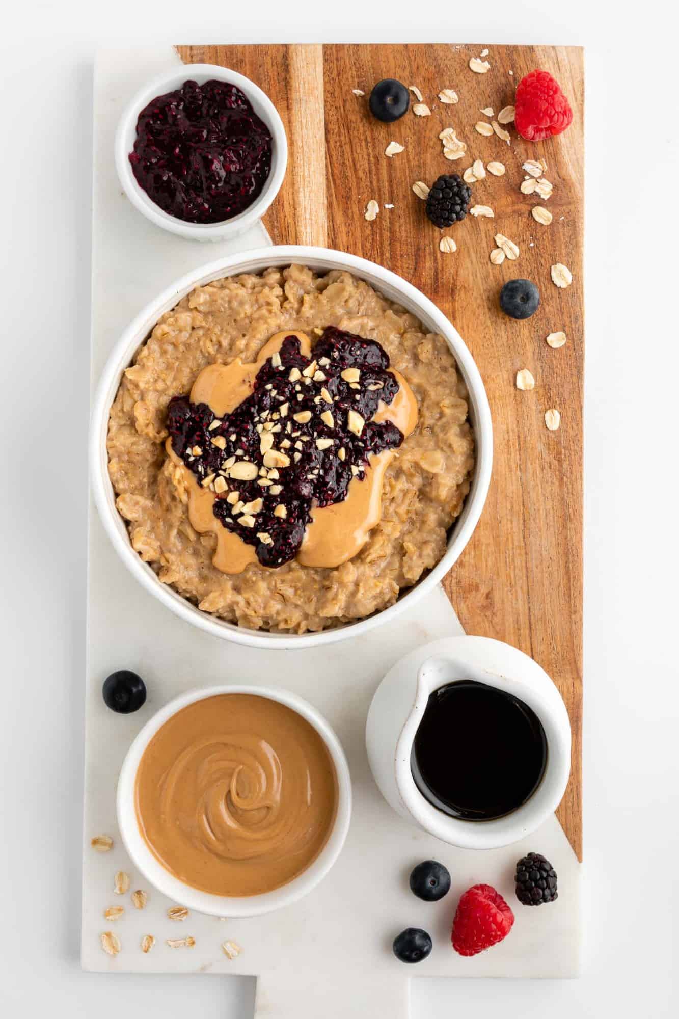 a wood and marble serving board topped with a bowl of pb&j oatmeal, a bowl of peanut butter, maple syrup, fresh berries, and oats