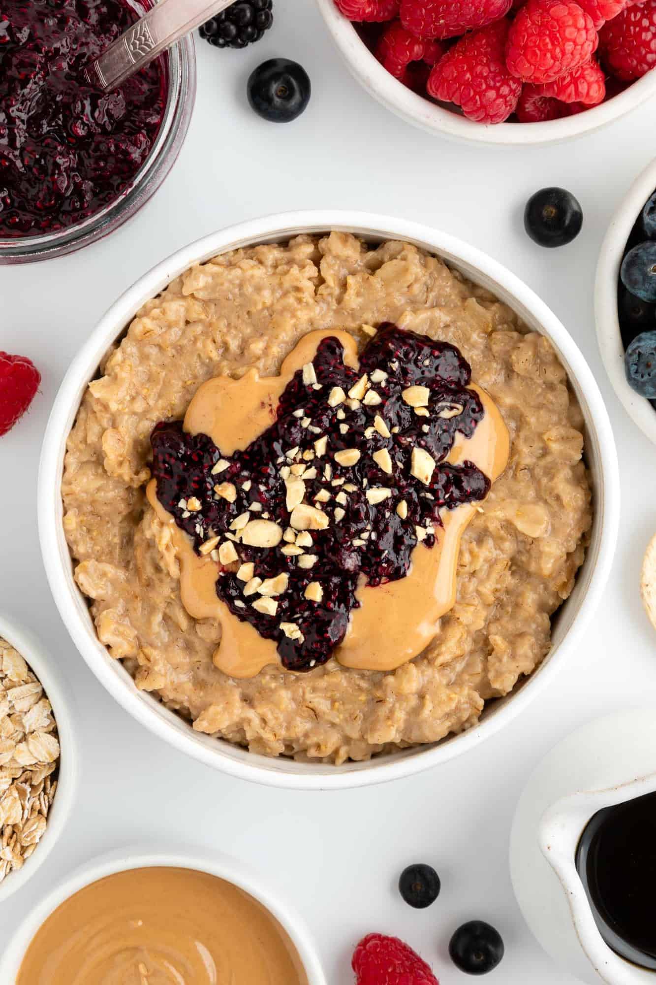 peanut butter and jelly inside a white bowl surrounded by berries, oats, maple syrup, and berry jam