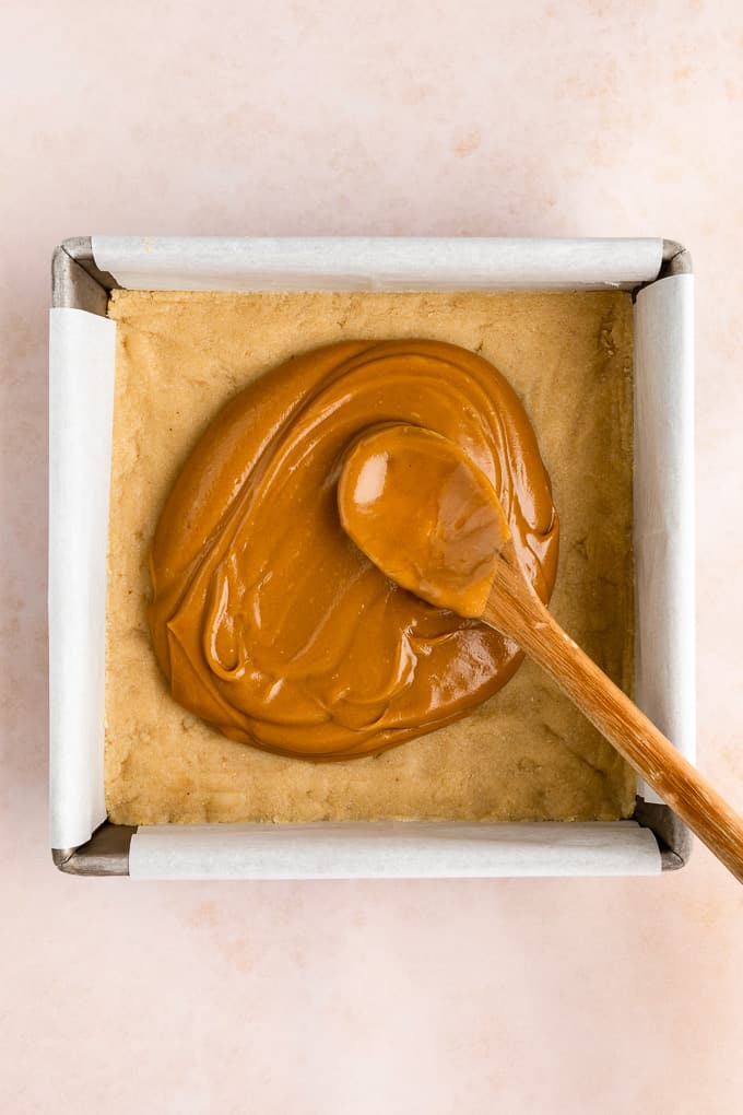 a wooden spoon spreading healthy vegan caramel across shortbread crust in a square baking dish