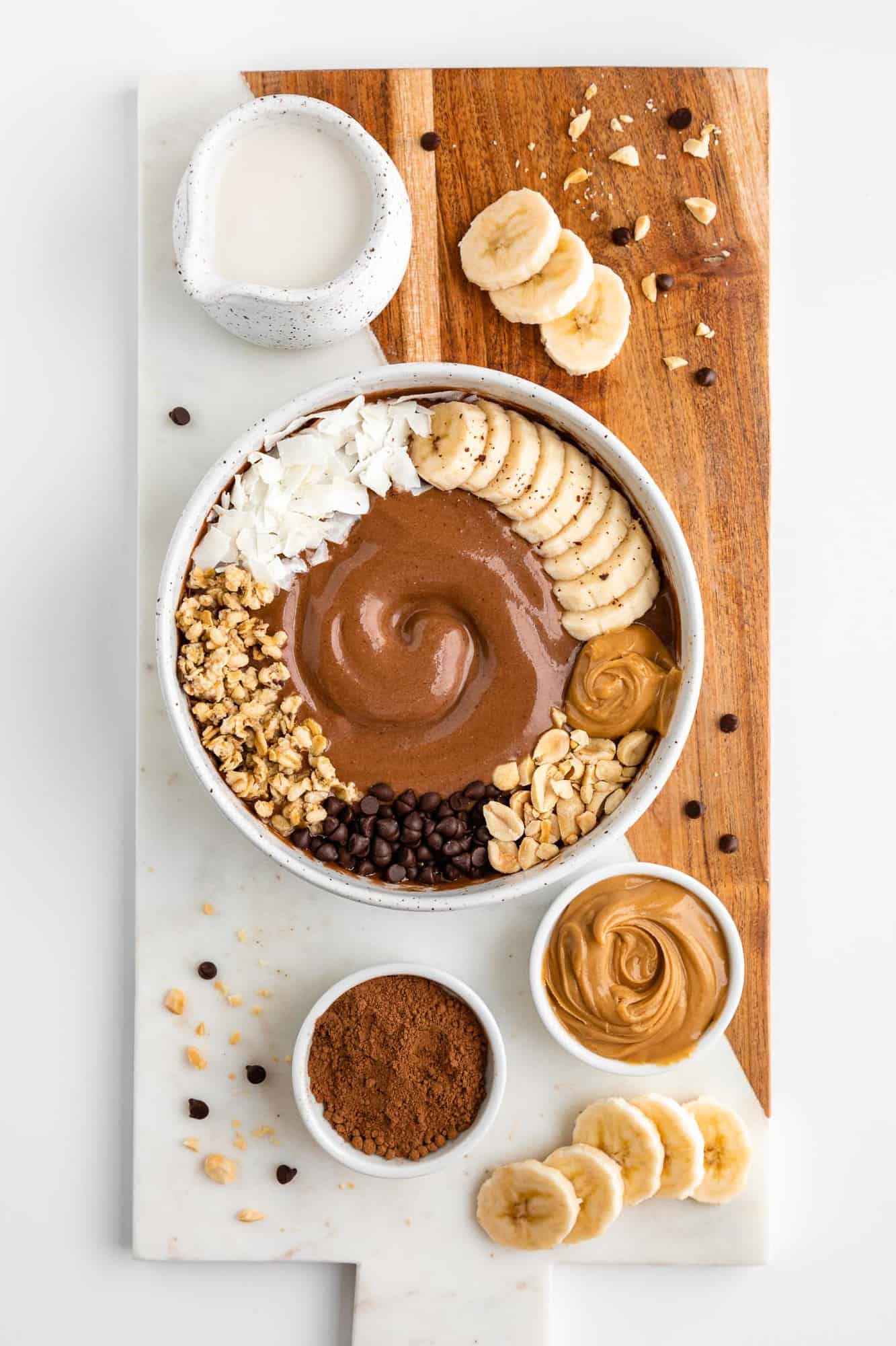 a wood and marble serving board topped with a chocolate peanut butter smoothie bowl surrounded by ingredients, including almond milk, cocoa powder, and banana