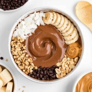 chocolate peanut butter smoothie bowl surrounded by bowls of ingredients, including banana, chocolate chips, and almond milk