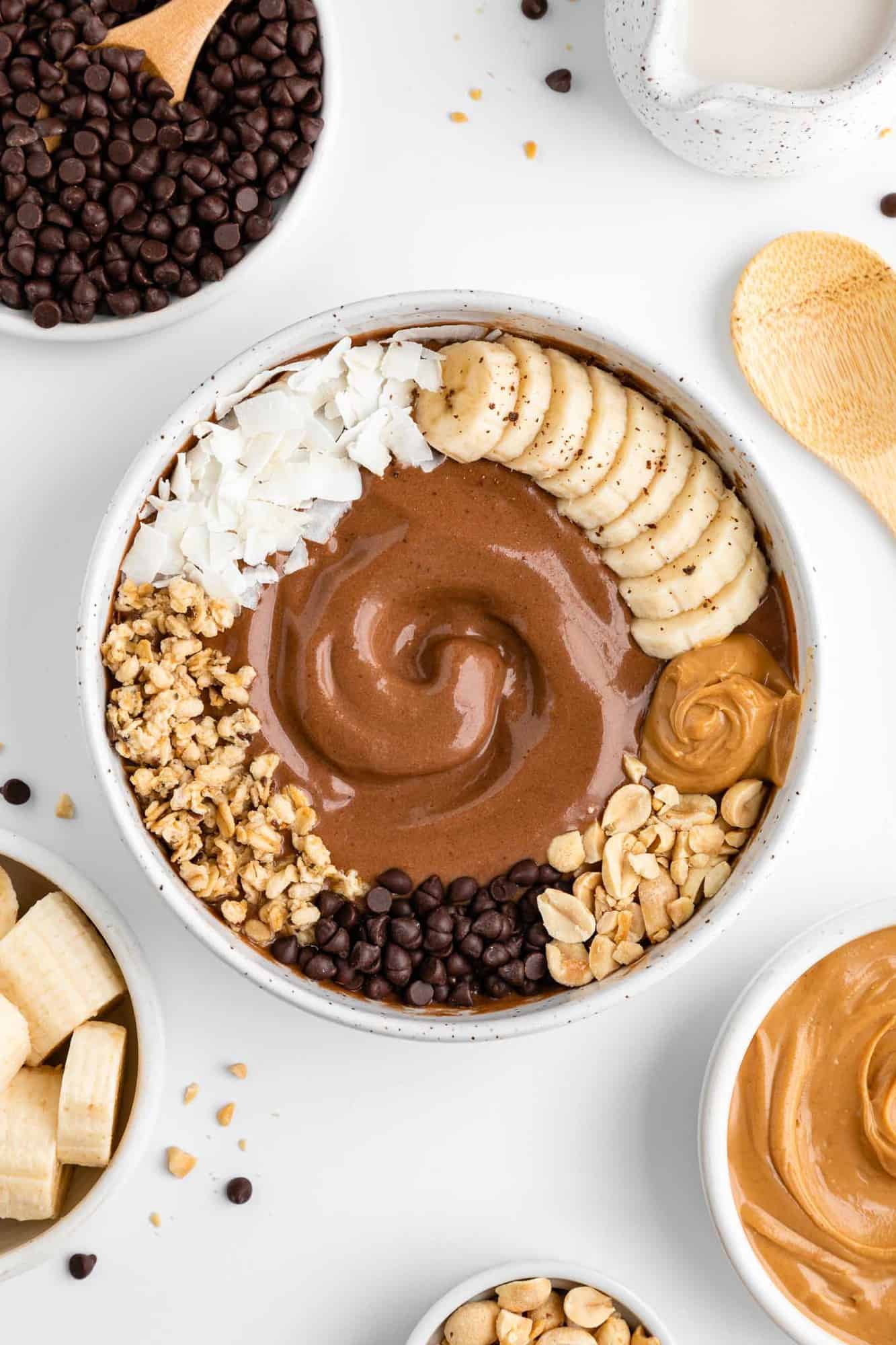 chocolate peanut butter smoothie bowl surrounded by bowls of ingredients, including banana, chocolate chips, and almond milk