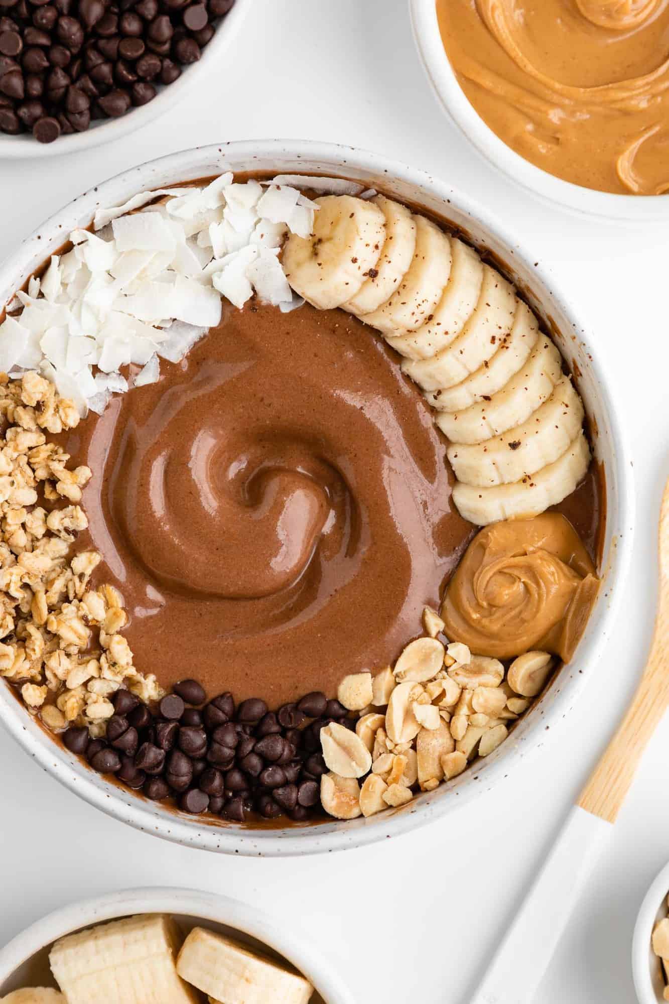 close up image of a chocolate peanut butter smoothie bowl surrounded by a bowl of bananas, a bamboo spoon, a bowl of nut butter, and a bowl of chocolate chips