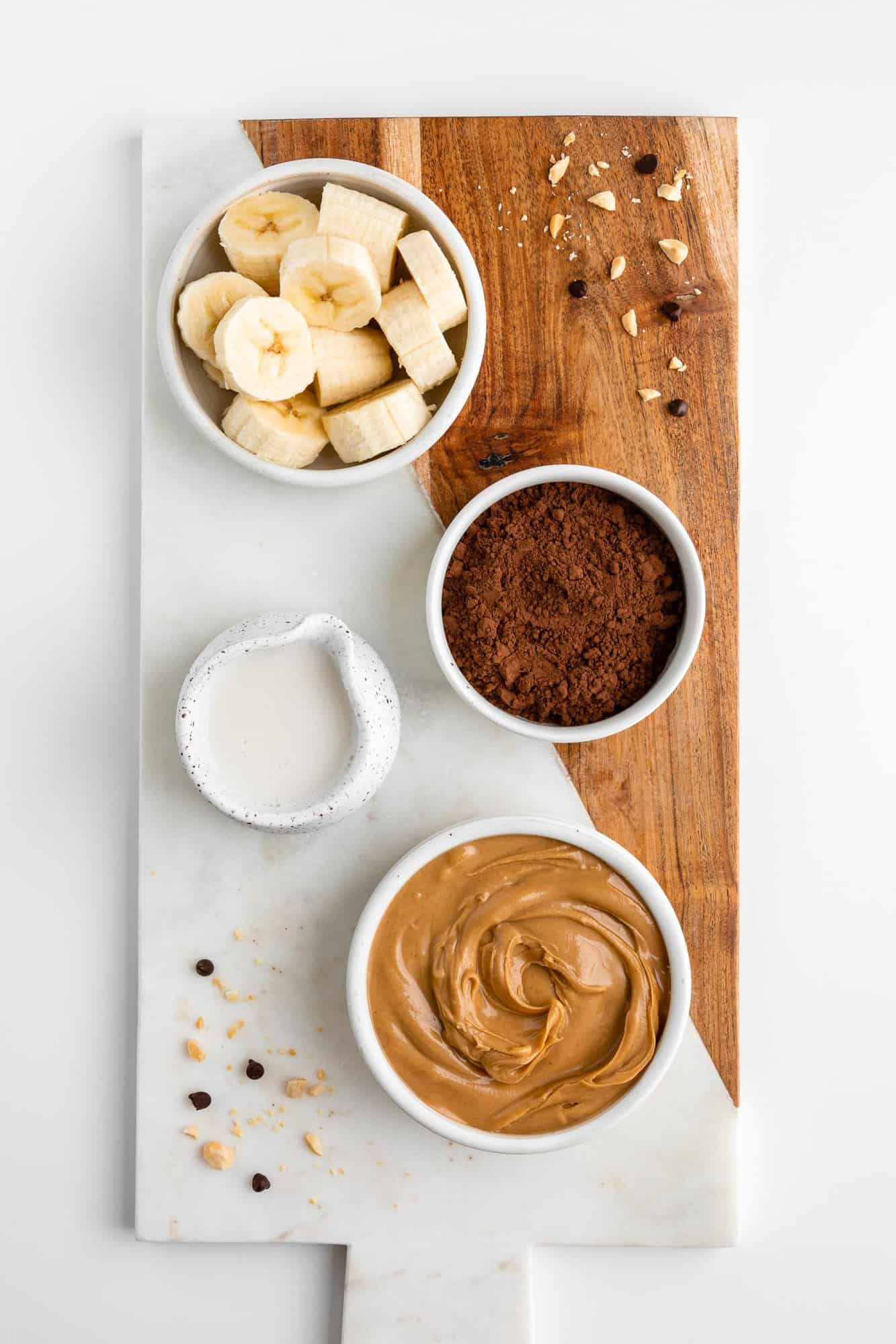 a wood and marble serving board topped with small bowls containing banana, cocoa powder, peanut butter, and almond milk