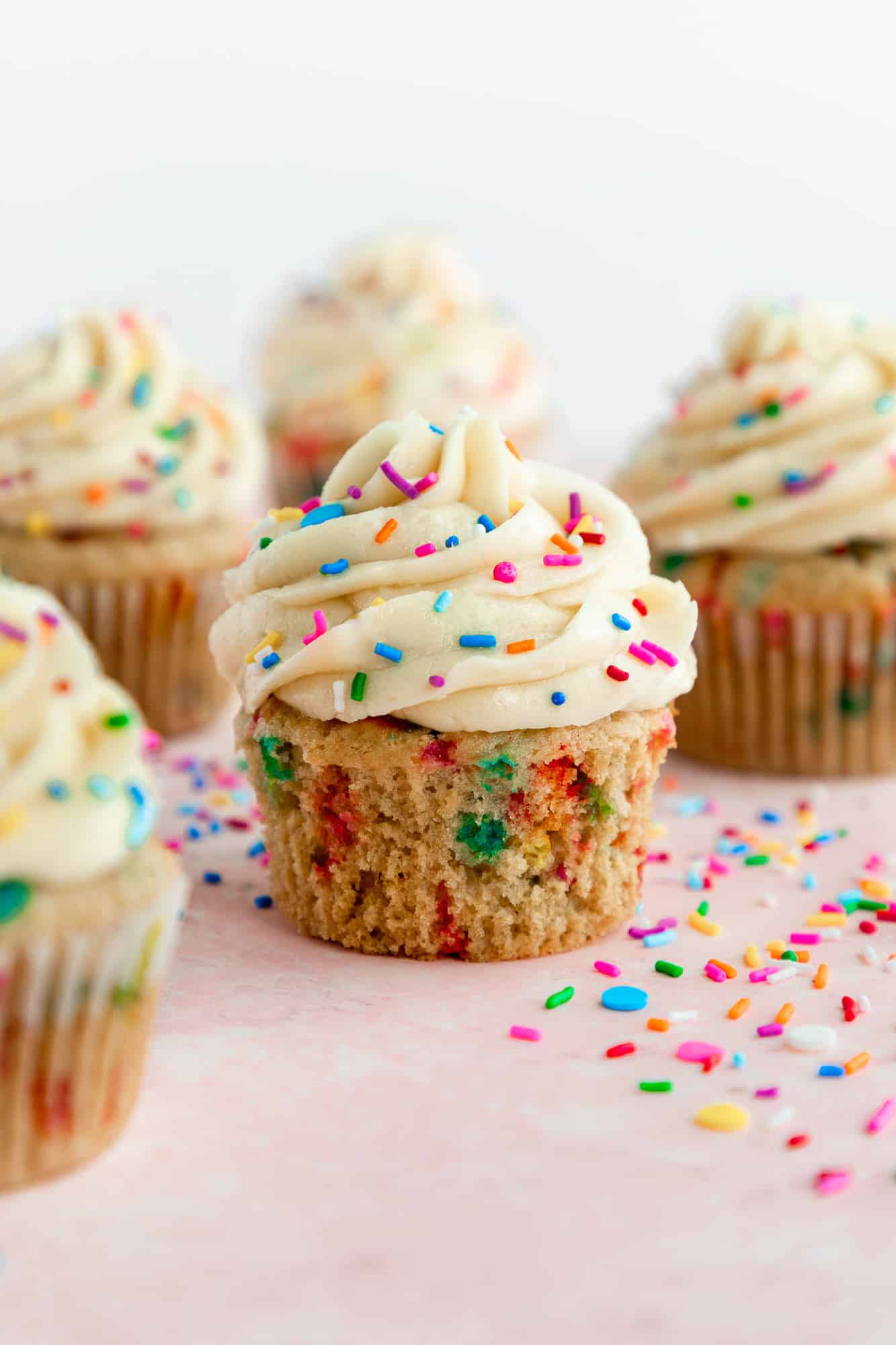 vegan funfetti cupcakes with vanilla frosting and rainbow sprinkles on a pink surface