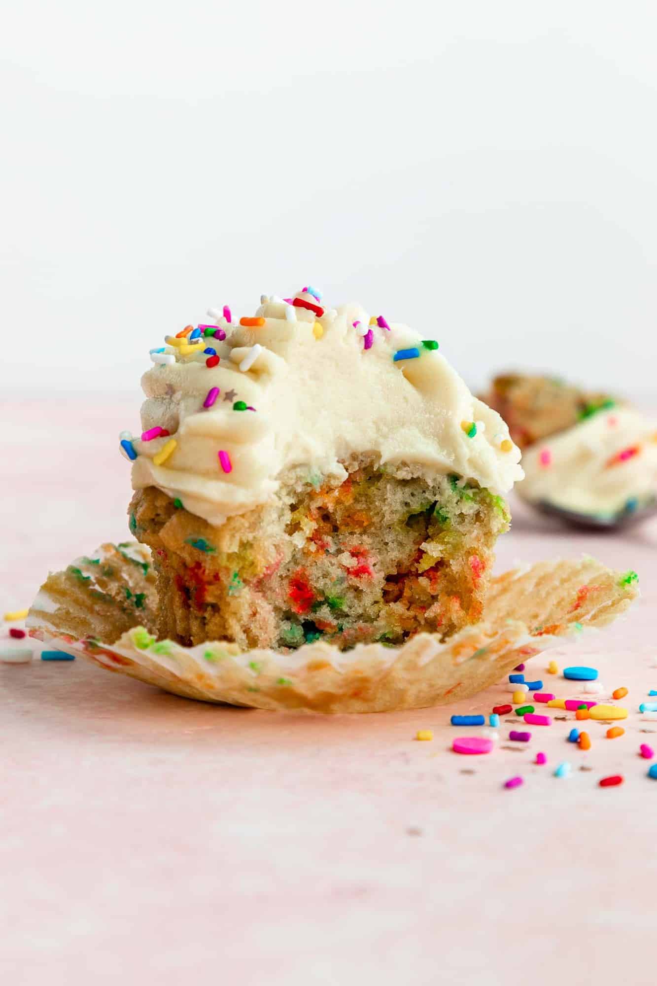 a partially eaten vegan funfetti cupcake topped with vanilla buttercream frosting and rainbow sprinkles