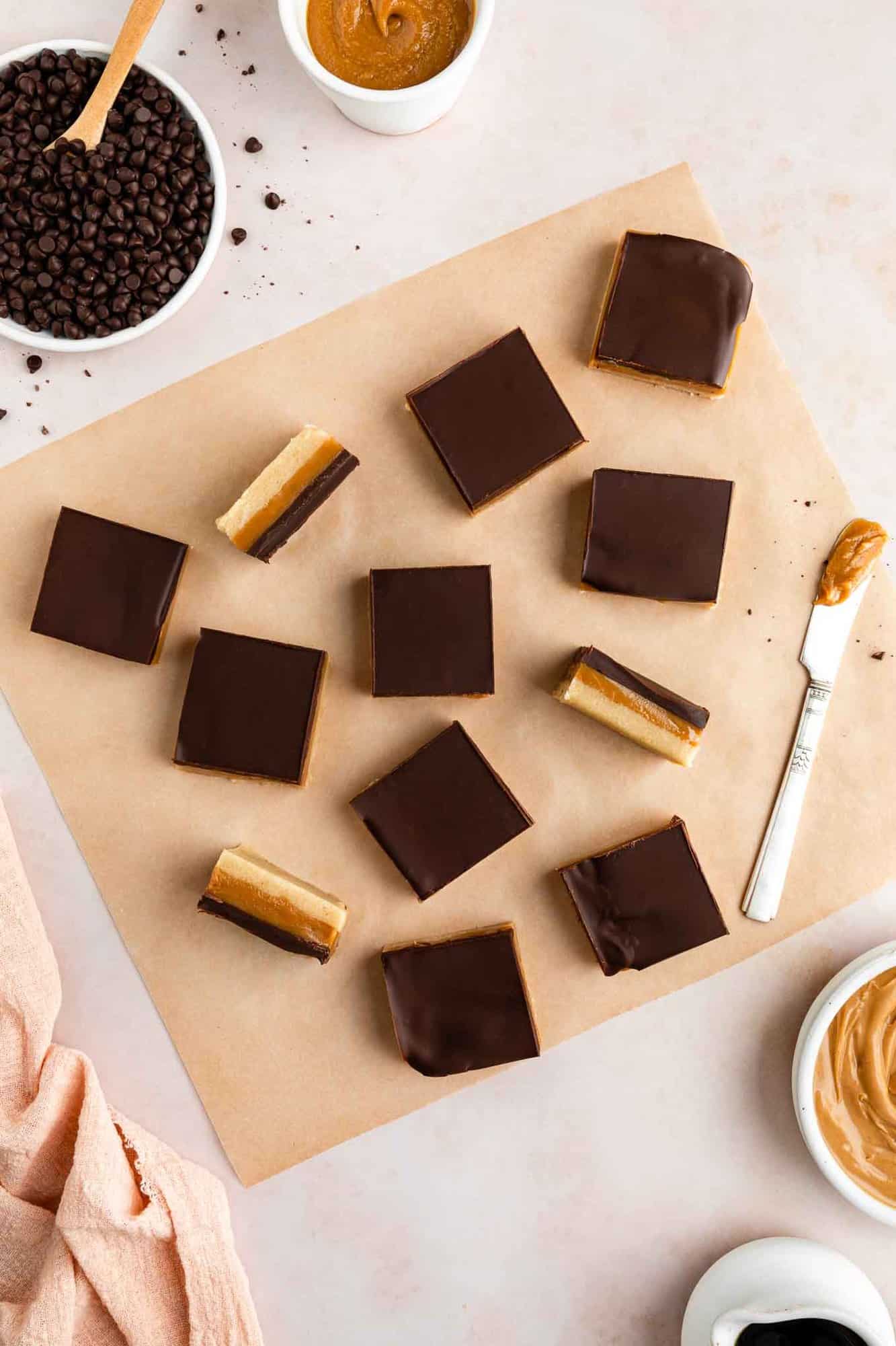 flatlay image of no-bake vegan millionaire shortbread bars on a square of brown parchment paper, surrounded by bowls of ingredients including chocolate chips and peanut butter