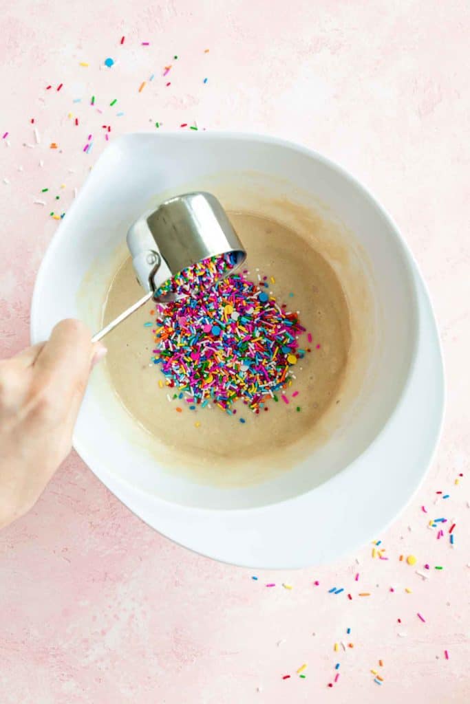 a hand pouring a measuring cup full of rainbow sprinkles into vegan funfetti cake batter