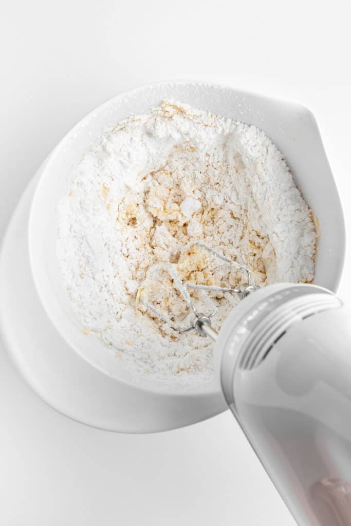 powdered sugar and vegan butter being mixed with a handheld electric mixer in a white bowl