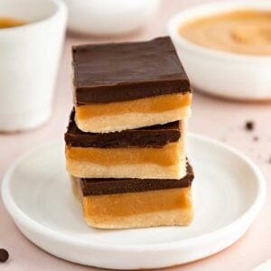 a stack of three no-bake vegan millionaire shortbread bars on a white plate beside a bowl of peanut butter and chocolate chips