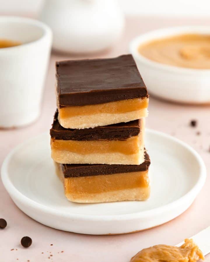 a stack of three no-bake vegan millionaire shortbread bars on a white plate beside a bowl of peanut butter and chocolate chips