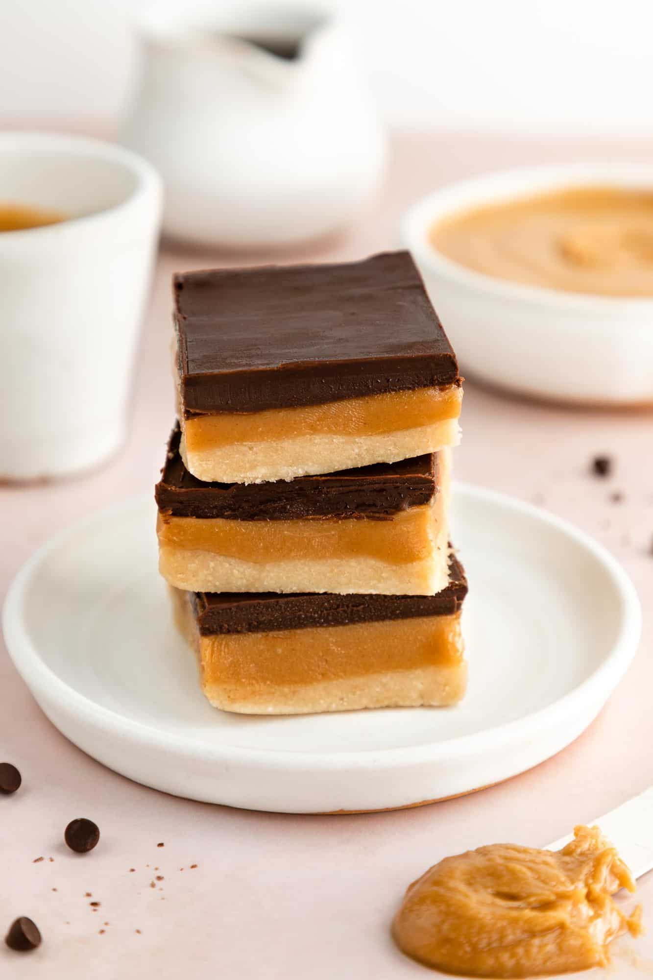 a stack of three no-bake vegan millionaire shortbread bars on a white plate beside a bowl of peanut butter and chocolate chips