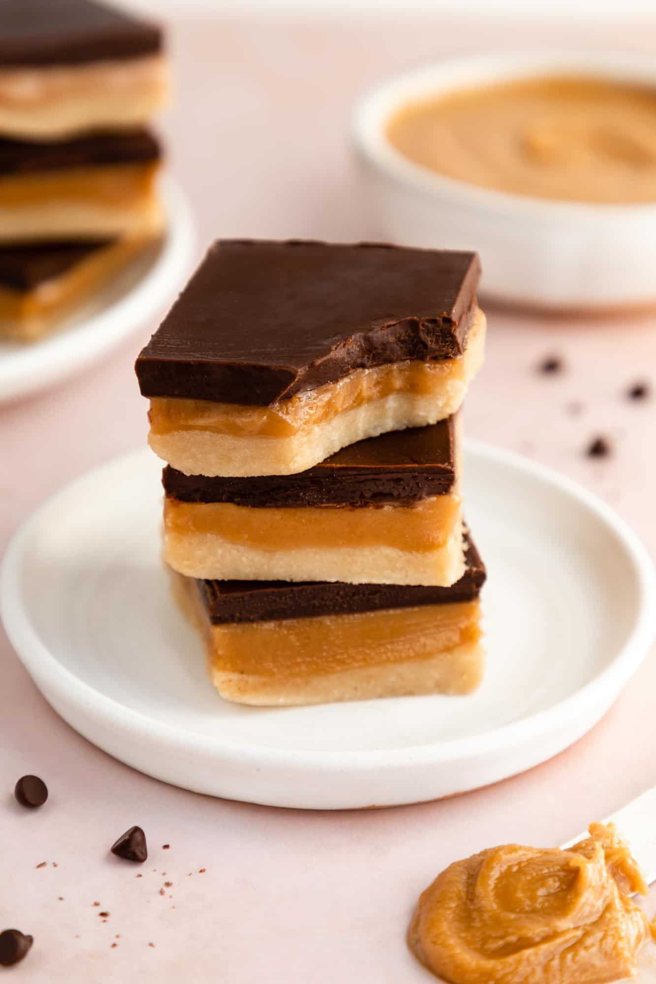 a stack of no-bake vegan millionaire shortbread bars on a white plate with a bite taken out of the top bar