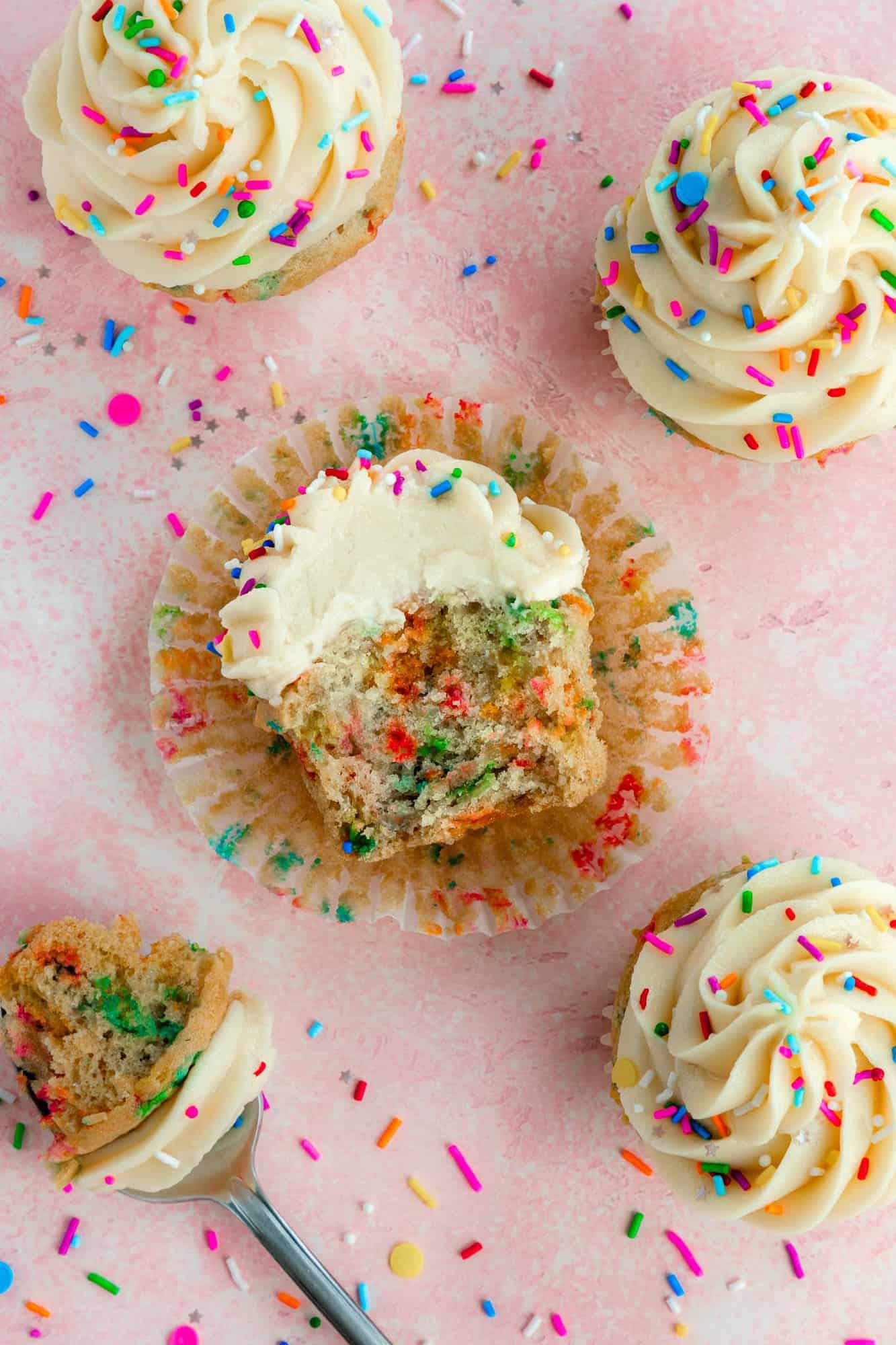 a partially eaten vegan funfetti cupcake topped with vanilla buttercream frosting and rainbow sprinkles laying on a peeled cupcake liner