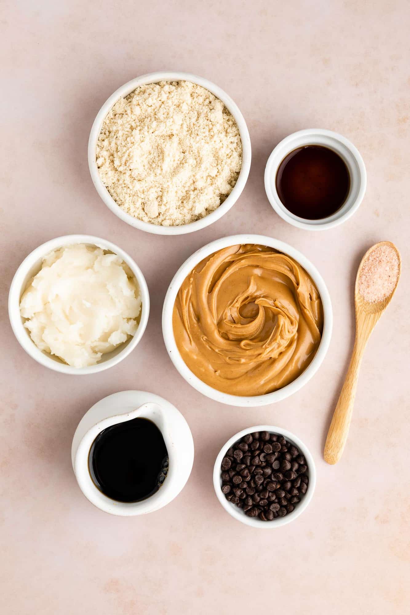 white bowls filled with peanut butter, almond flour, coconut oil, maple syrup, chocolate chips, and vanilla extract on a blush colored backdrop