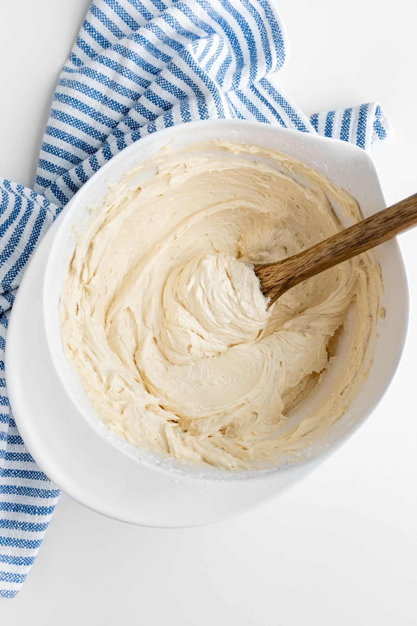 a wooden spoon mixing vegan vanilla frosting in a white mixing bowl beside a blue striped kitchen towel