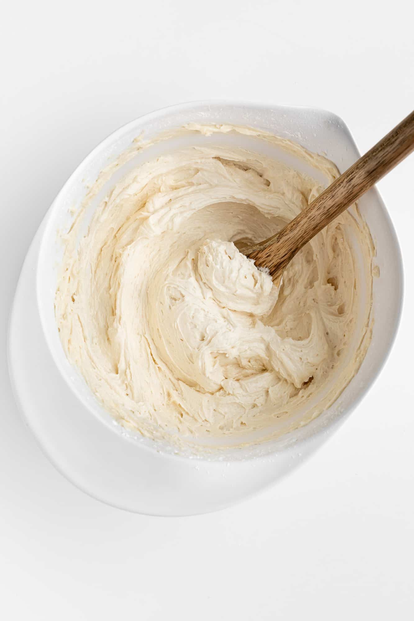a wooden spoon mixing vegan vanilla frosting in a white mixing bowl