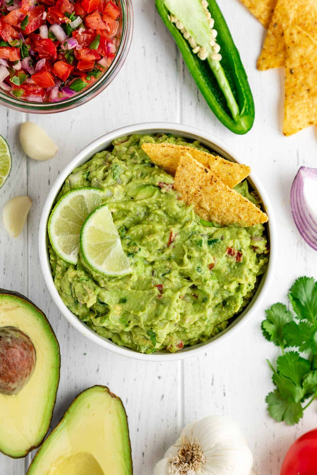 best guacamole in a white bowl with corn tortilla chips, limes, and surrounded by guac ingredients