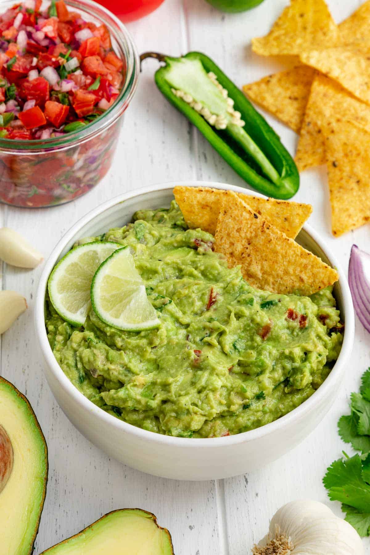 best guacamole in a white bowl with corn tortilla chips, limes, and surrounded by guac ingredients