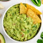 corn tortilla chips inside a bowl of guacamole surrounded by guac ingredients