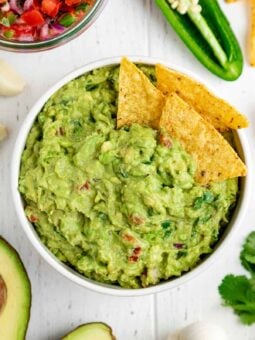 corn tortilla chips inside a bowl of guacamole surrounded by guac ingredients