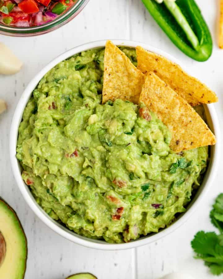 corn tortilla chips inside a bowl of guacamole surrounded by guac ingredients
