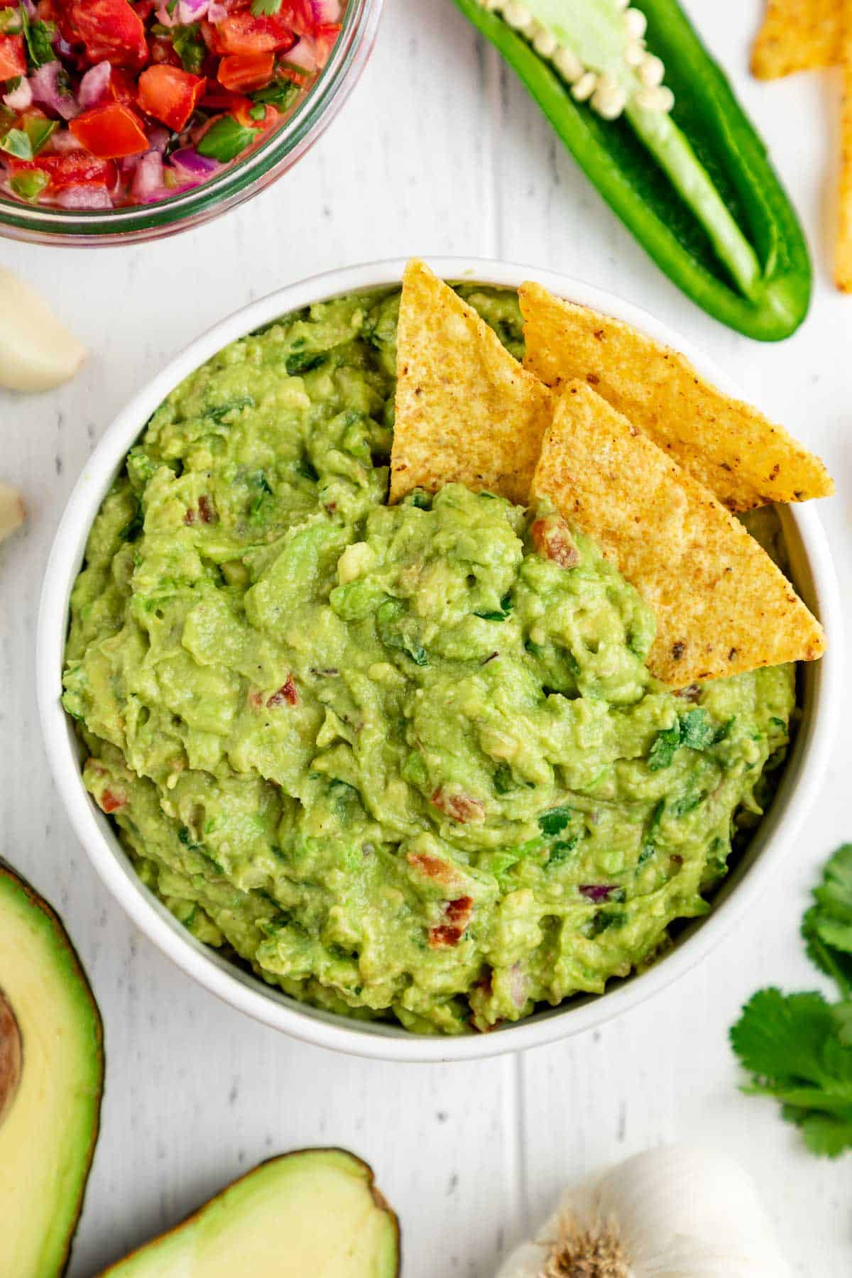 corn tortilla chips inside a bowl of guacamole surrounded by guac ingredients