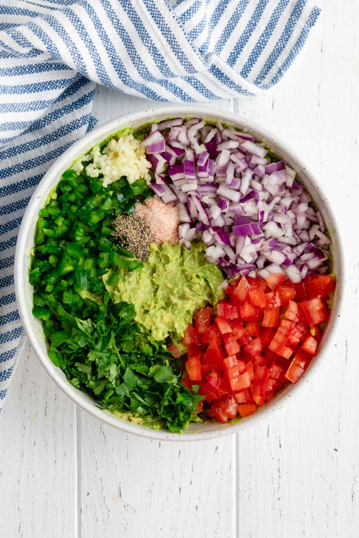 a white bowl filled with mashed avocado, diced tomato, red onion, cilantro, jalapeño, garlic, salt, and pepper