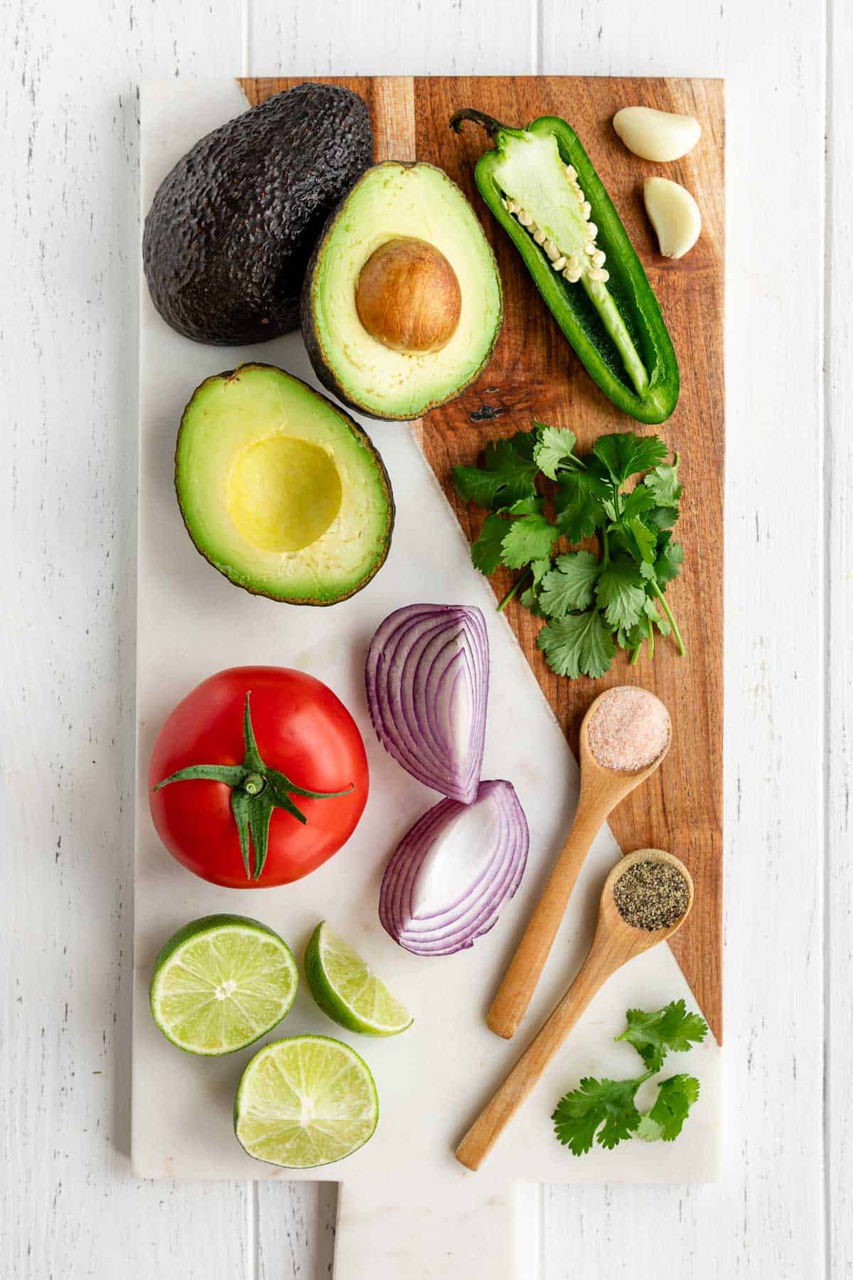 a marble serving board topped with avocado, tomato, red onion, limes, cilantro, jalapeño, garlic, salt, and pepper
