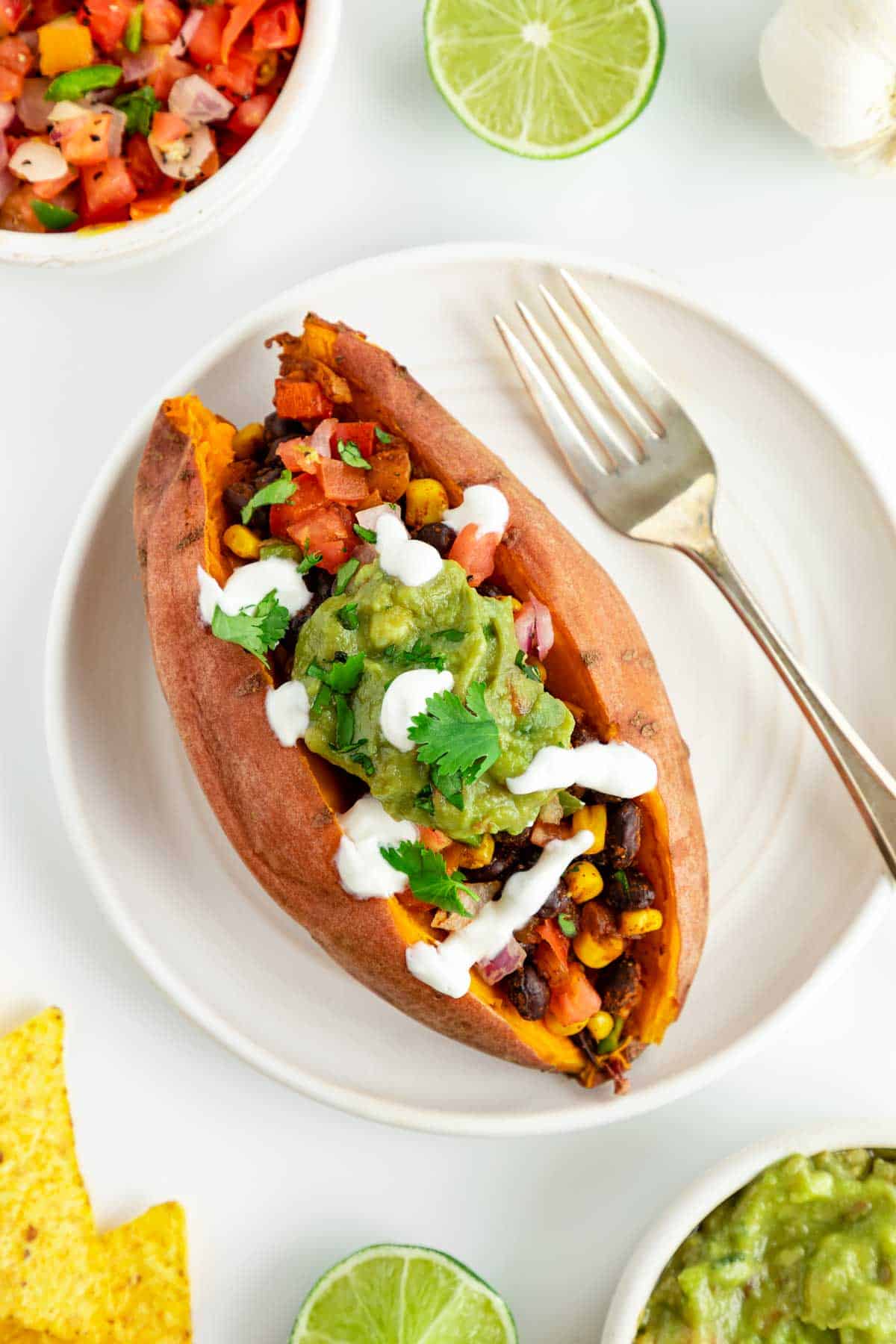 a mexican stuffed sweet potato on a white plate surrounded by bowls of guacamole, pico de gallo, and corn tortilla chips