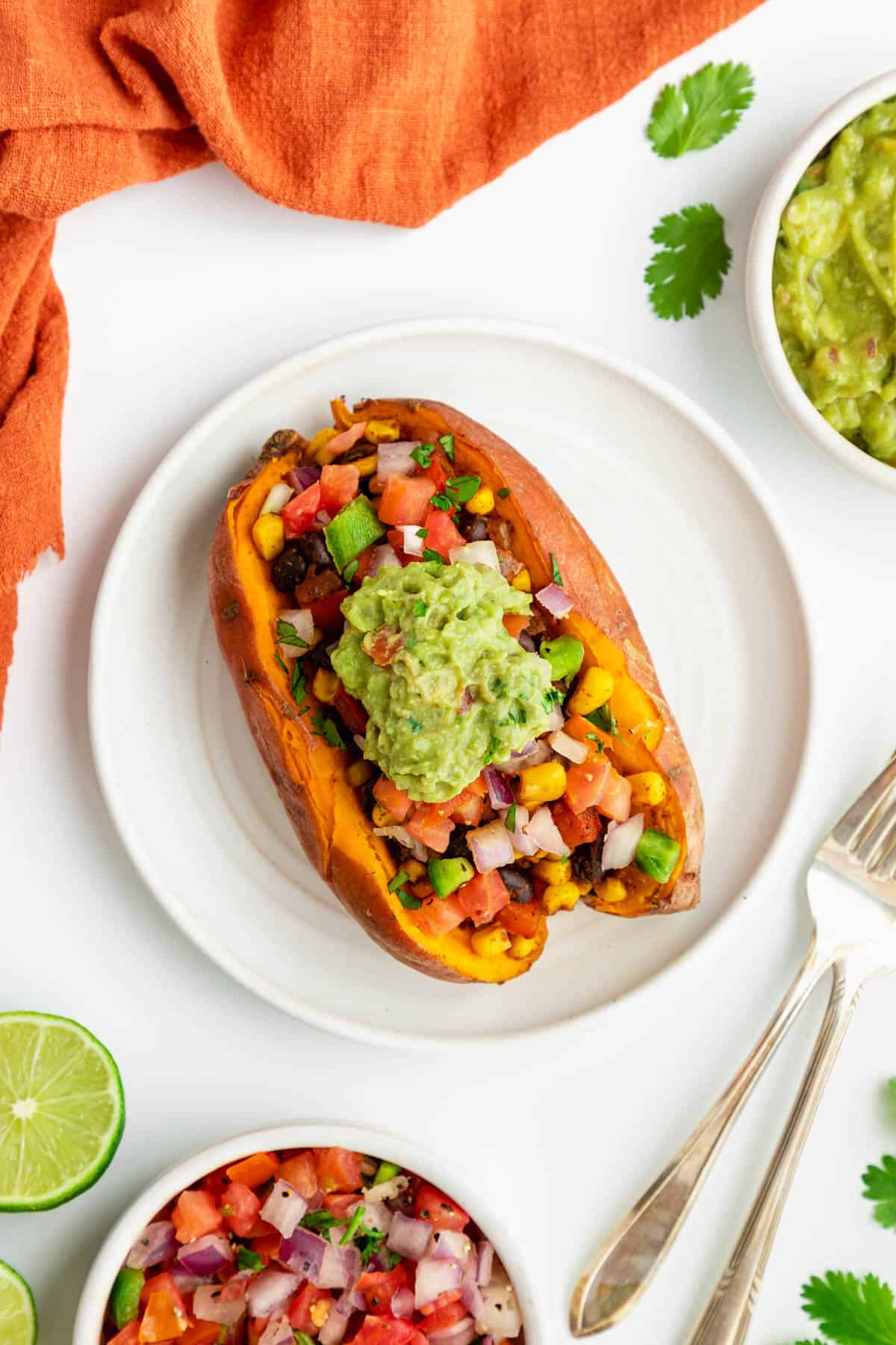 a sweet potato stuffed with black beans, corn, and guacamole on a white plate surrounded by ingredients