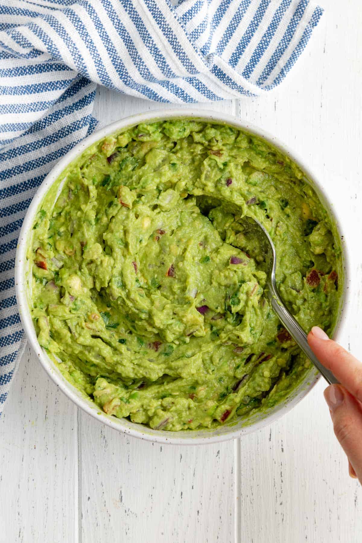 a hand mixing guacamole in a white bowl beside a striped kitchen towel