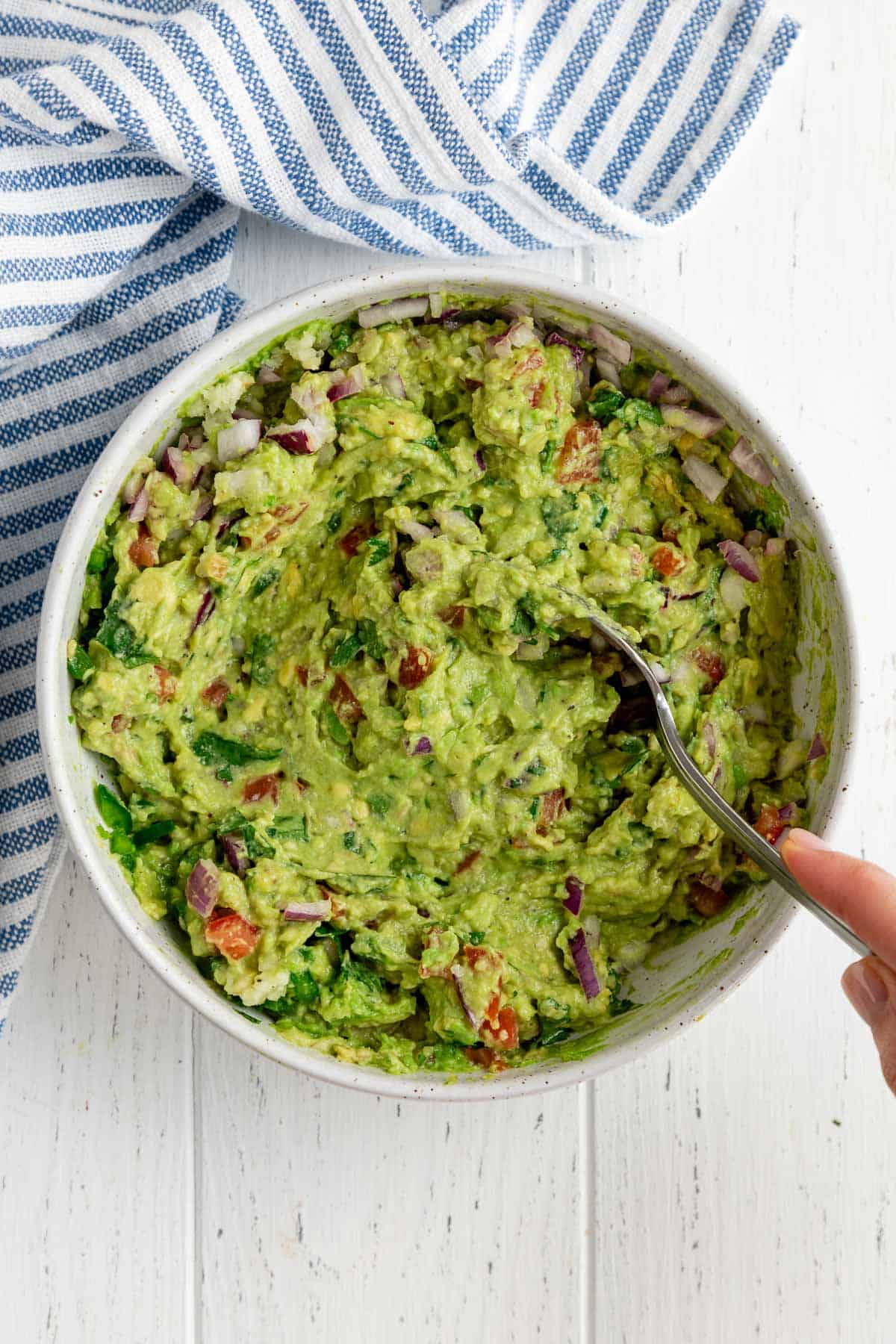 hand mixing guacamole in a large white bowl beside a striped kitchen towel