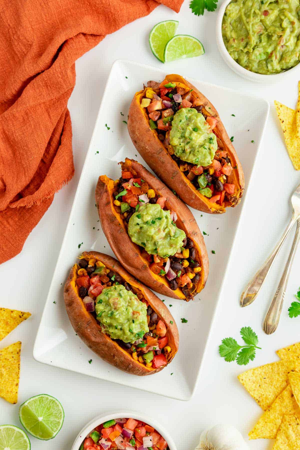 three mexican stuffed sweet potatoes on a white rectangular plate surrounded by corn tortilla chips, guacamole, limes, pico de gallo, and an orange linen napkin