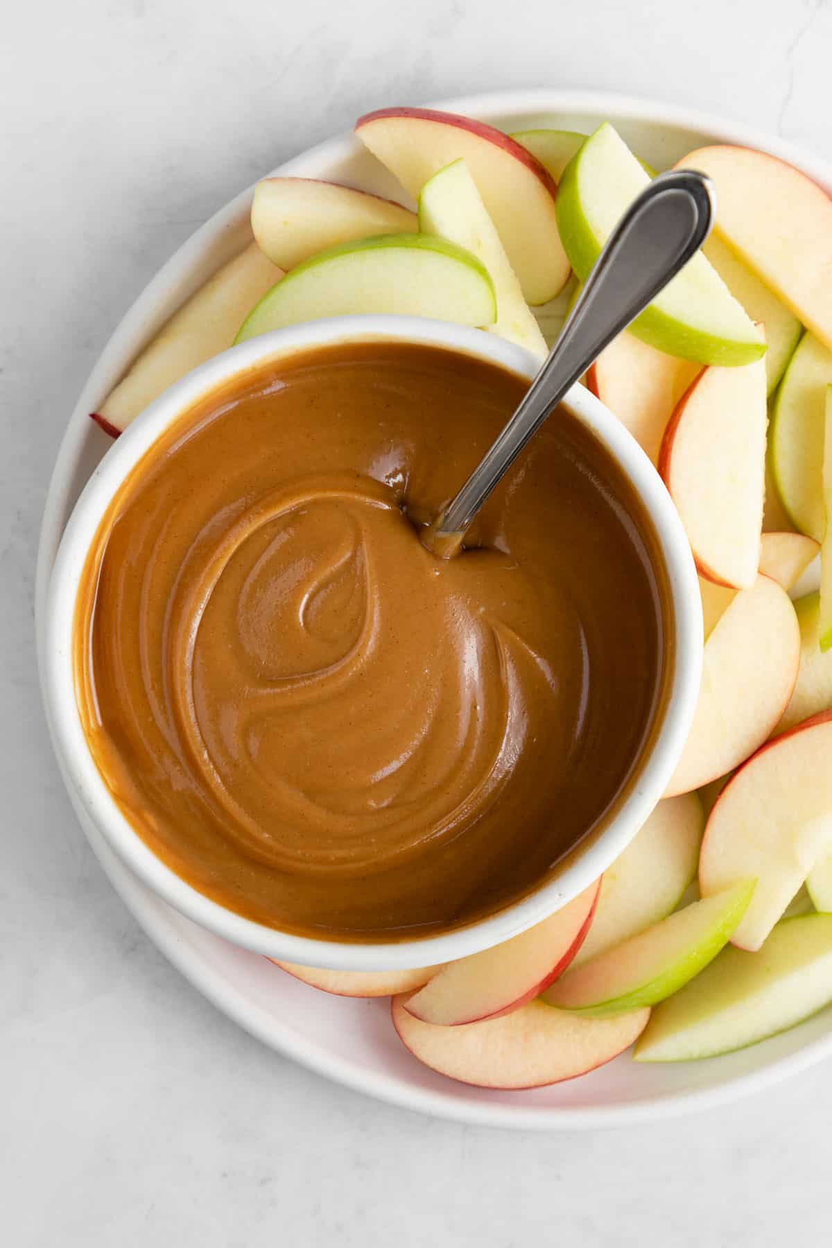 healthy caramel dip in a white bowl with a spoon surrounded by apple slices