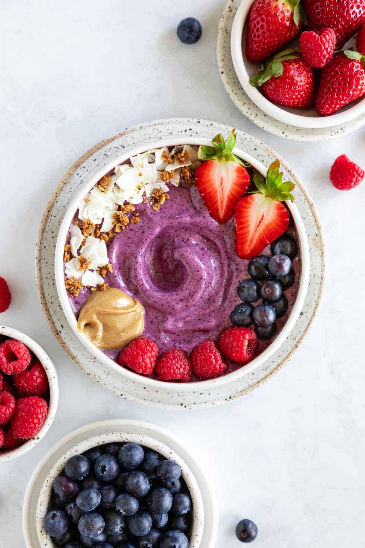 berry smoothie bowl on a speckled ceramic plate surrounded by bowls of blueberries, raspberries, and strawberries