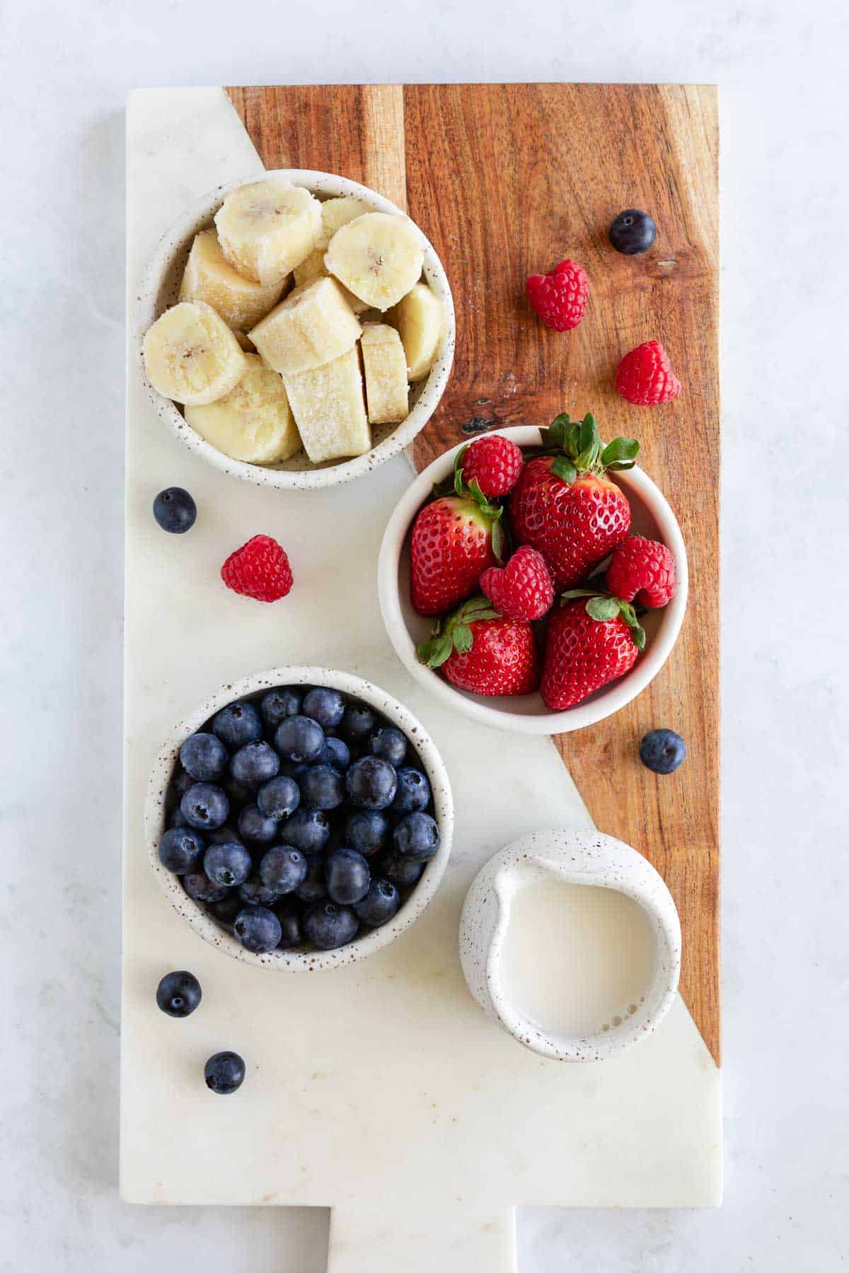 a marble and wood serving board topped with small ceramic bowls filled with frozen bananas, strawberries, blueberries, and almond milk