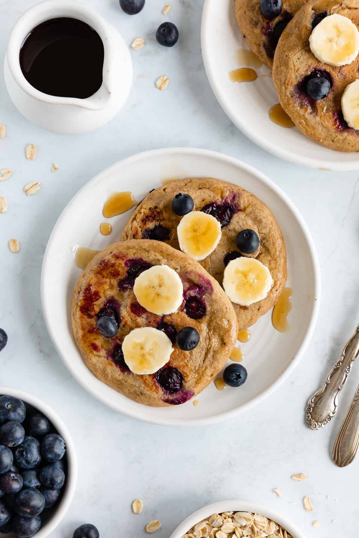 vegan blueberry banana pancakes on two white plates beside a pitcher of maple syrup