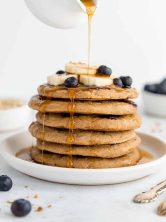 maple syrup being poured over a stack of vegan gluten-free blueberry banana pancakes