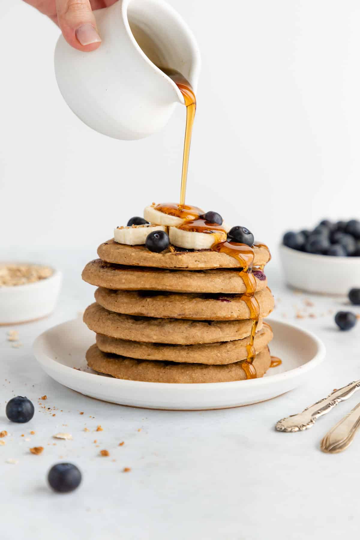 a hand pouring maple syrup over a stack of vegan blueberry banana pancakes